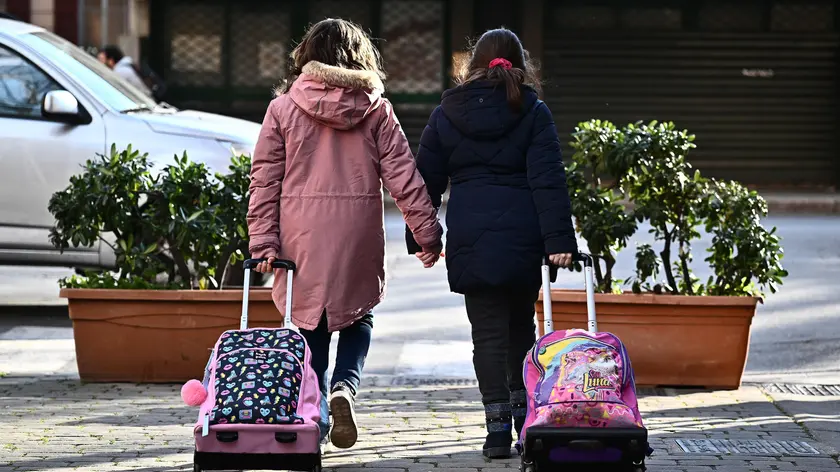 Due bambine si tengono la mano mentre vanno a scuola trascinando i loro zaini a Genova, in una foto d'archivio del 3 febbraio 2023. ANSA/LUCA ZENNARO (scuola, bambini, bambine, educazione, istruzione, simbolica, simboliche, generica, zaino, libri, studio, cultura, infanzia)