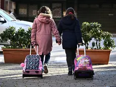 Due bambine si tengono la mano mentre vanno a scuola trascinando i loro zaini a Genova, in una foto d'archivio del 3 febbraio 2023. ANSA/LUCA ZENNARO (scuola, bambini, bambine, educazione, istruzione, simbolica, simboliche, generica, zaino, libri, studio, cultura, infanzia)