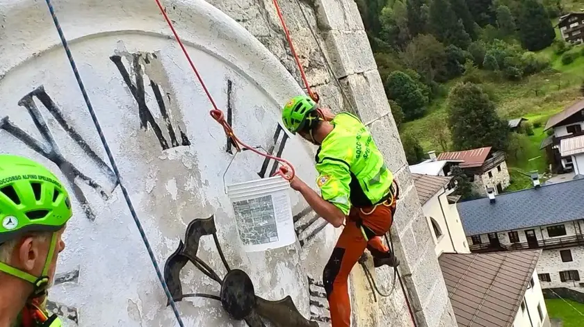 I tecnici della Stazione del Soccorso alpino di Forni di Sopra, imbragati con corde e funi, all’opera per ridipingere l’orologio che guarda a Sud del campanile della chiesa nella frazione di Cella