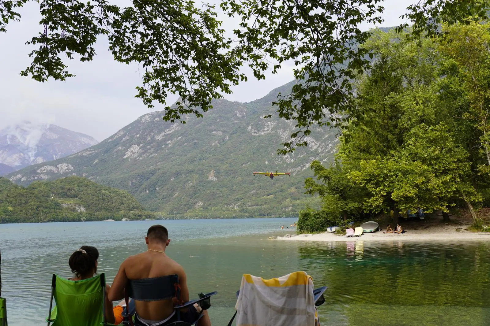 I turisti lungo le sponde del lago di Cavazzo mentre il canadair raccoglie l'acqua per spegnere gli incendi