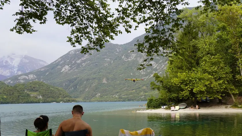 I turisti lungo le sponde del lago di Cavazzo mentre il canadair raccoglie l'acqua per spegnere gli incendi