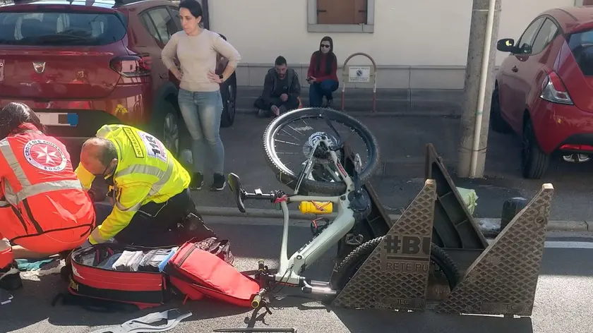 I soccorsi al ciclista caduto e, in primo piano, la bicicletta incastrata nel tombino